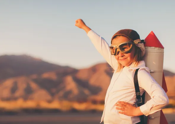 A child in a white shirt with a rucksack that looks like a rocket stands with his fist raised in front of a mountain landscape.