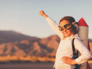 Ein Kind in einem weißen Hemd mit einem Rucksack, der wie eine Rakete aussieht, steht mit erhobener Faust vor einer Berglandschaft.