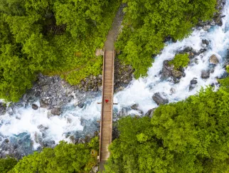 Luftaufnahme einer hölzernen Brücke über einen reißenden Fluss, umgeben von dichtem grünem Wald.