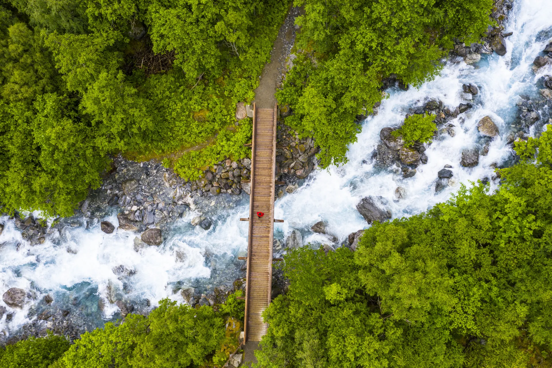 Luftaufnahme einer hölzernen Brücke über einen reißenden Fluss, umgeben von dichtem grünem Wald.